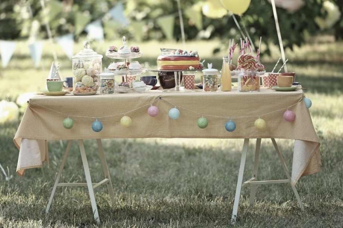 Gartentisch, der mit einer Tischdecke bedeckt ist und an dem eine Geburtstagstisch gedeckt ist. Auf dem Tisch steht ein Geburtstagskuchen, Süßikeiten und getränle. Die Vorderseite ist mit einer Girlande aus runden Bällen geschmückt