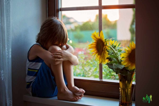 Kleinkind sitzt traurig auf der Fensterbank  und hat die Knie angezogen und die Armerum herum geschlungen. Links von ihm ist ein Stück durchsichtige Gardine, rechts steht eine Blumenvase mit Sonnenblumen